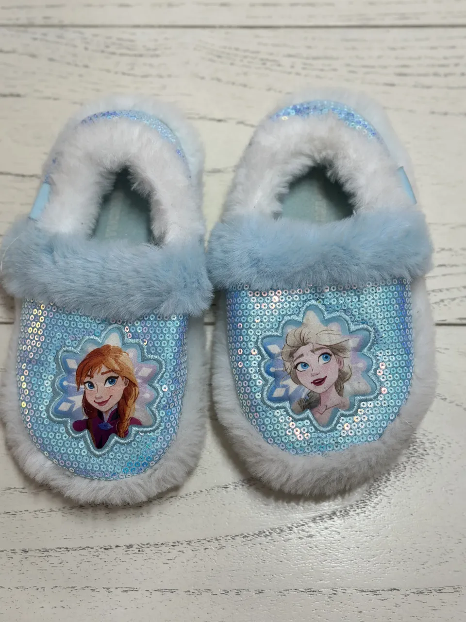 Pantuflas de niña con personajes de Frozen, Anna y Elsa. Diseño con lentejuelas celestes y detalles de peluche blanco y celeste.