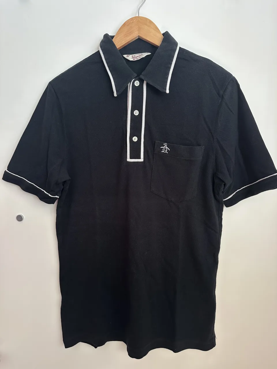 Remera manga corta negra con cuello polo y detalles de ribb blanco en cuello, puños y tapacostura de botones. Posee un bolsillo en el pecho con un bordado de pingüino.