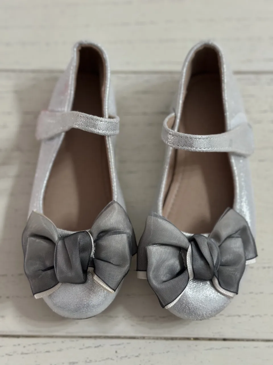 Zapatillas plateadas con brillos y moño gris en la punta. Tienen una tira con velcro para ajustar.