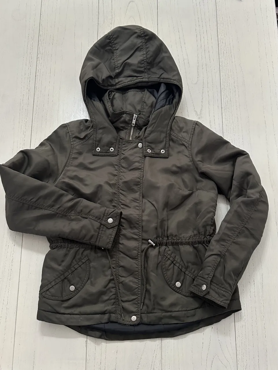 Campera con capucha, cierre y cordón ajustable en la cintura. Posee bolsillos con botones a presión.