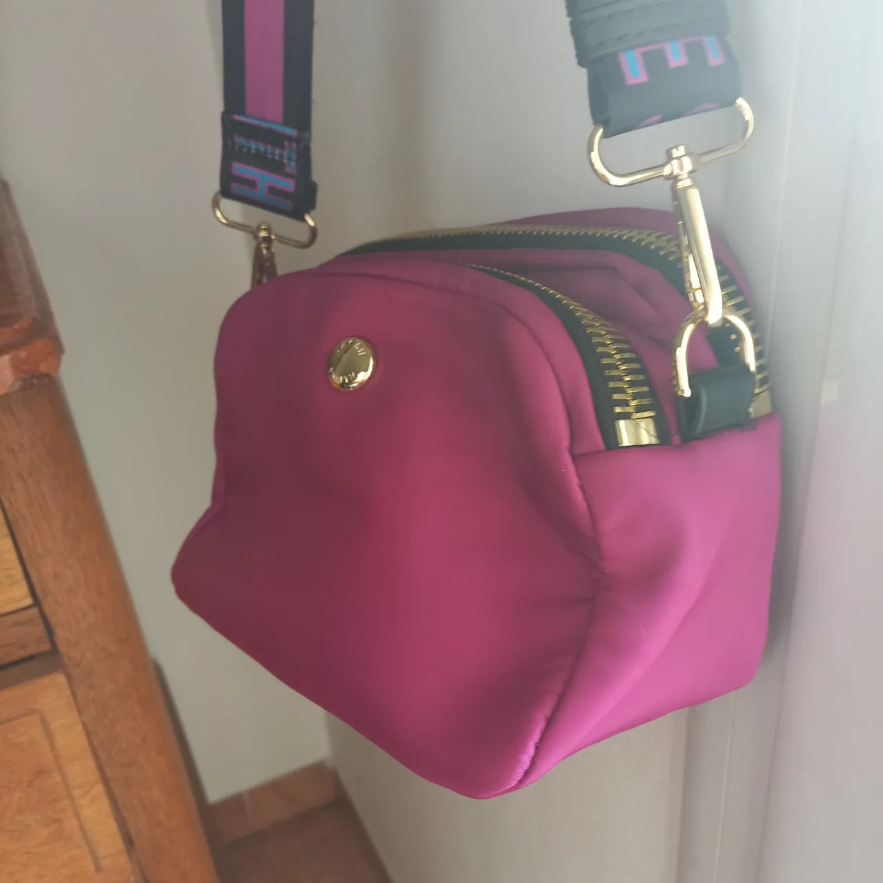 Cartera de color fucsia con dos cierres principales y correa ajustable con logo.