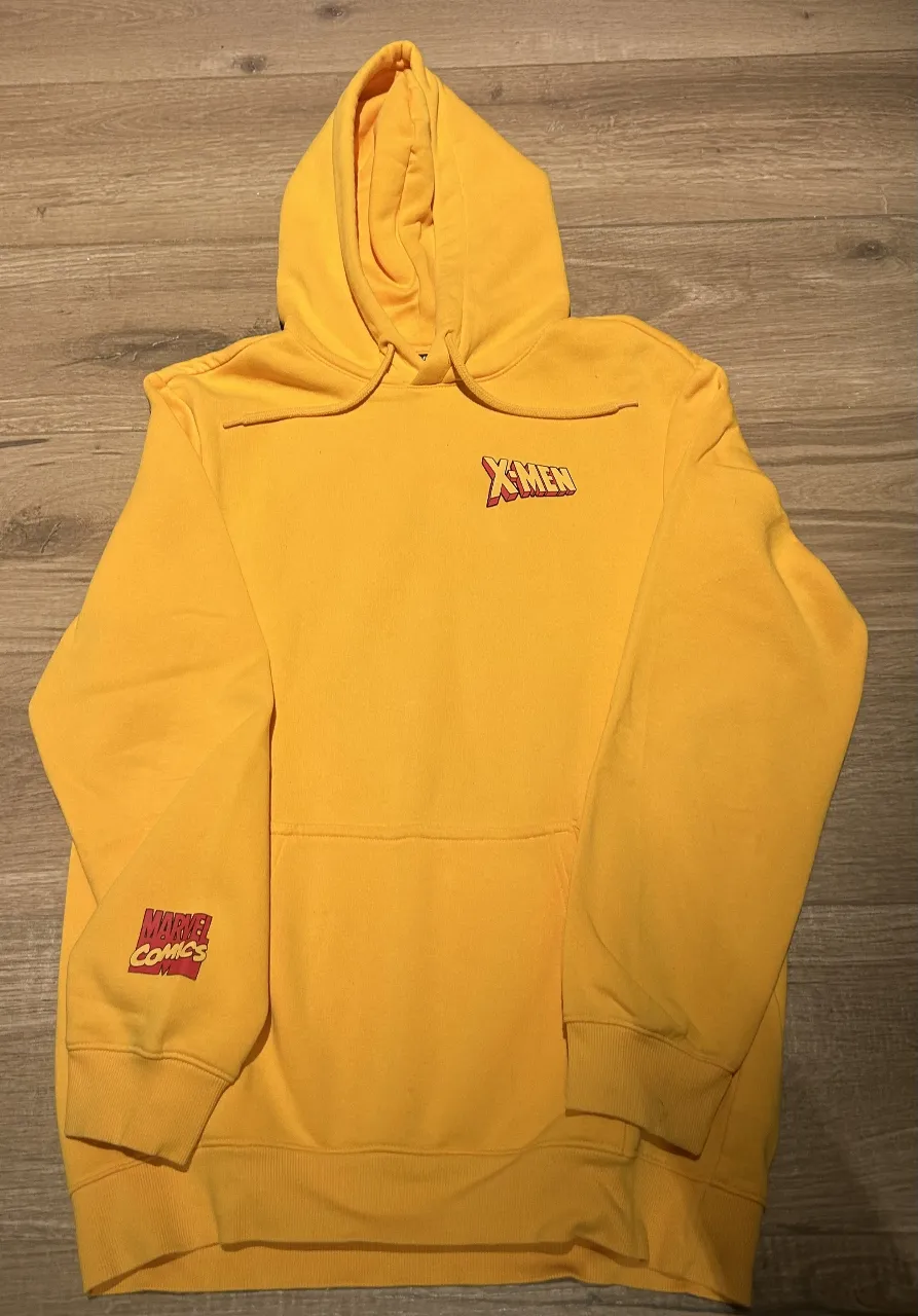 Buzo canguro amarillo de hombre con capucha y estampado de X-Men en la espalda. Tiene el logo de Marvel Comics en la manga.