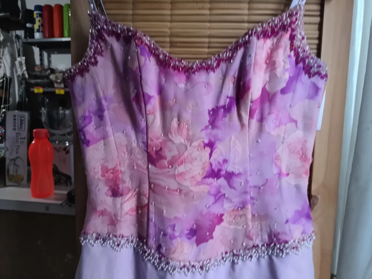Vestido largo con tiras finas. El corpiño es de seda estampada con flores en tonos lilas, rosas y fucsias, y está adornado con canutillos y perlas. La falda es lisa, de seda, en color lila claro y tiene un detalle de volado asimétrico.