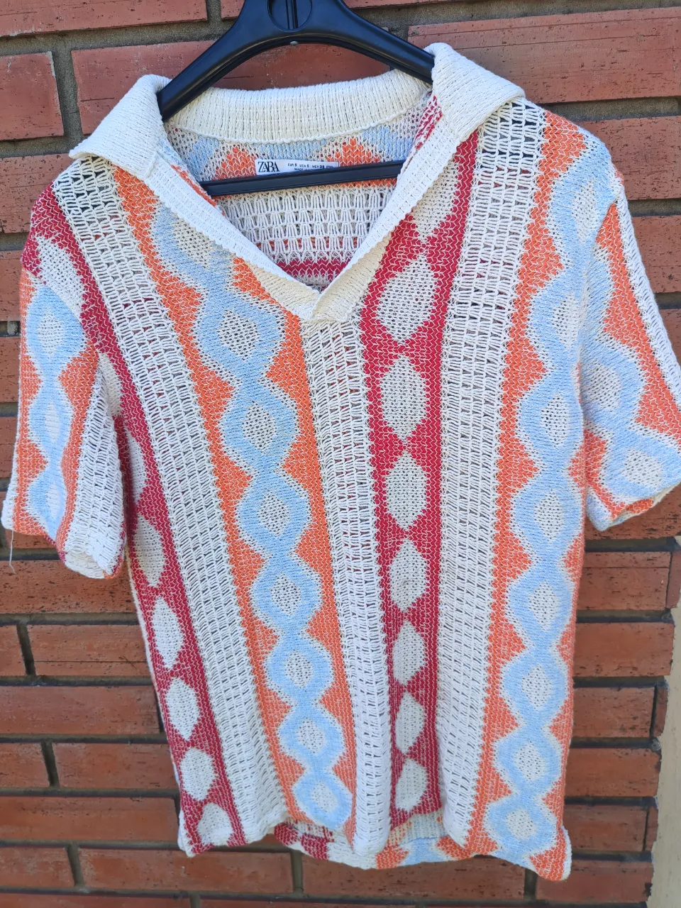 Remera de tejido de punto con cuello escote en V y mangas cortas. Presenta un estampado geométrico de rombos y líneas verticales en colores naranja, rojo, azul y blanco.