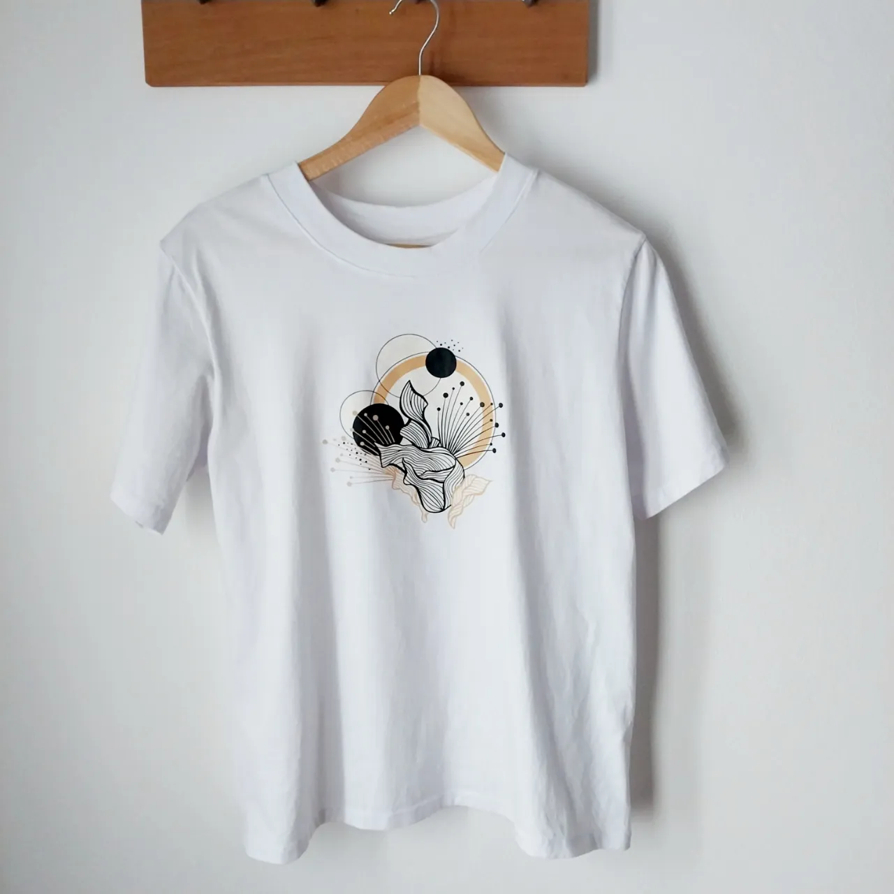 Remera blanca con estampa abstracta de flores y círculos en tonos negro, dorado y beige. Diseño moderno y original, ideal para un look casual y artístico.