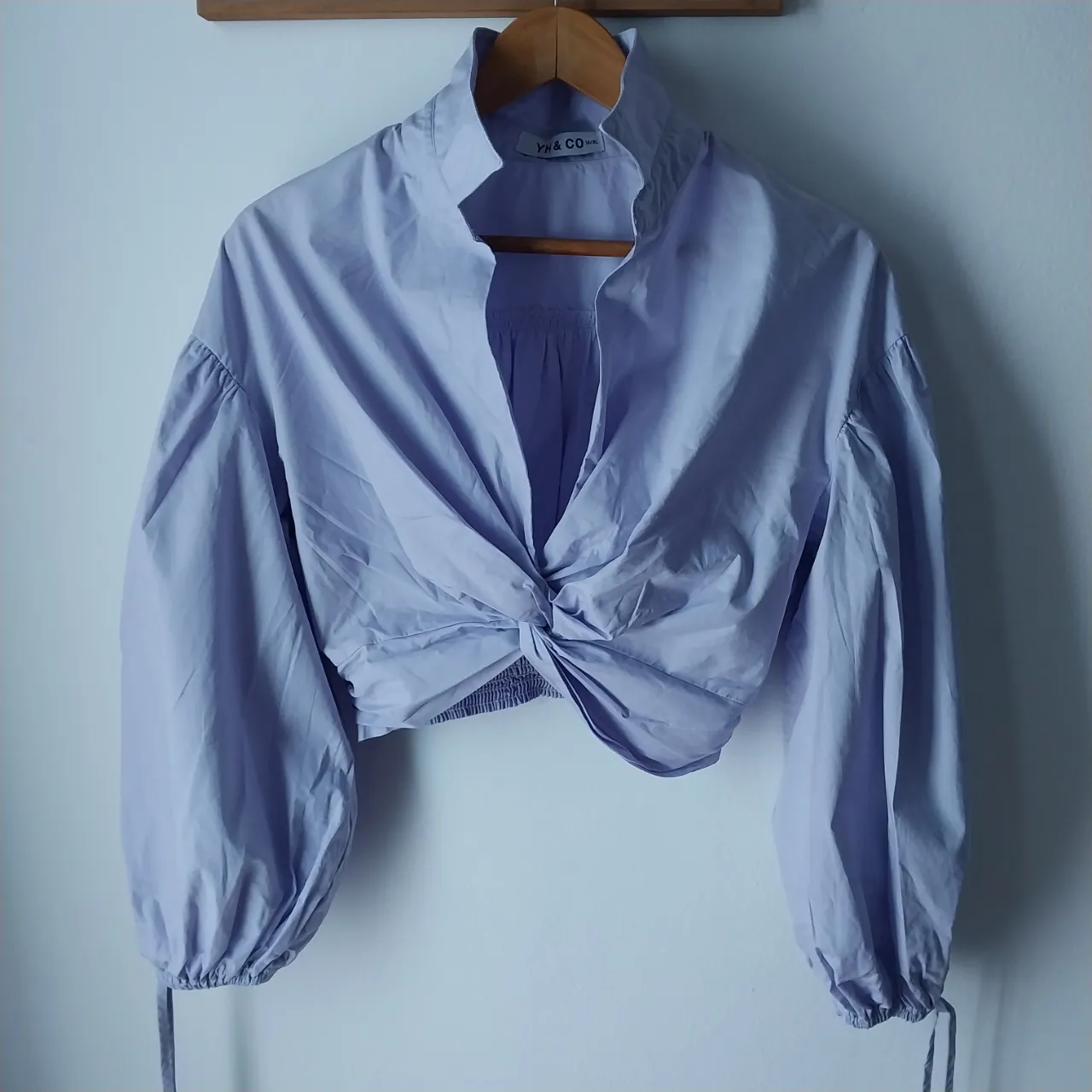 Blusa crop de popelina color lavanda, con cuello mao, mangas largas abullonadas con puños ajustables y detalle de nudo delantero. Un diseño moderno y chic, ideal para looks de día o noche.