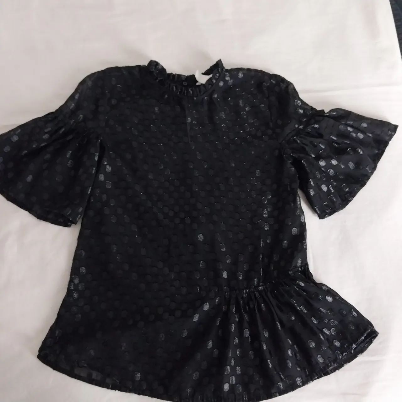 Blusa negra con mangas acampanadas y detalles de lunares brillantes. Posee un cuello mao con volados y un ruedo con fruncido que le da un toque femenino y moderno. Ideal para un look elegante y sofisticado.medidas sin estirar,hombros 36cm de axila a axila 43cm largo tomado desde hombro:62cm. mangas29cm.