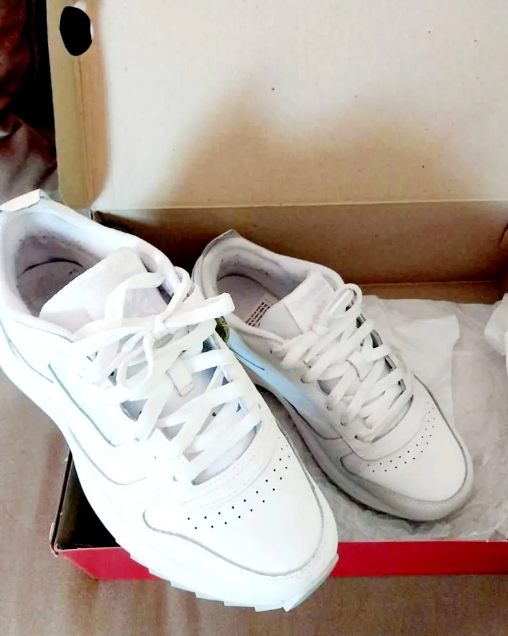 Zapatillas deportivas blancas con cordones y detalles grises. Tienen perforaciones en la punta y el logo de la marca en los laterales.