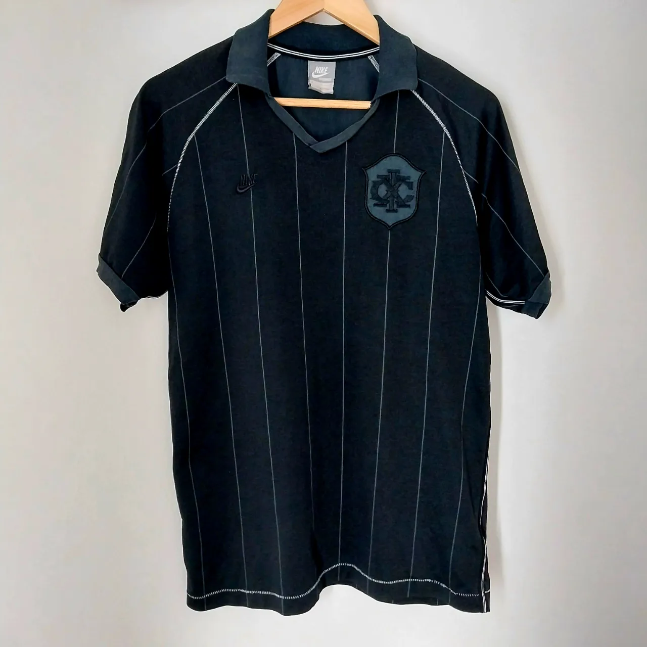 Remera deportiva Nike de color negro con finas rayas verticales grises. Posee cuello en V con solapa y logo de la marca bordado en el pecho.