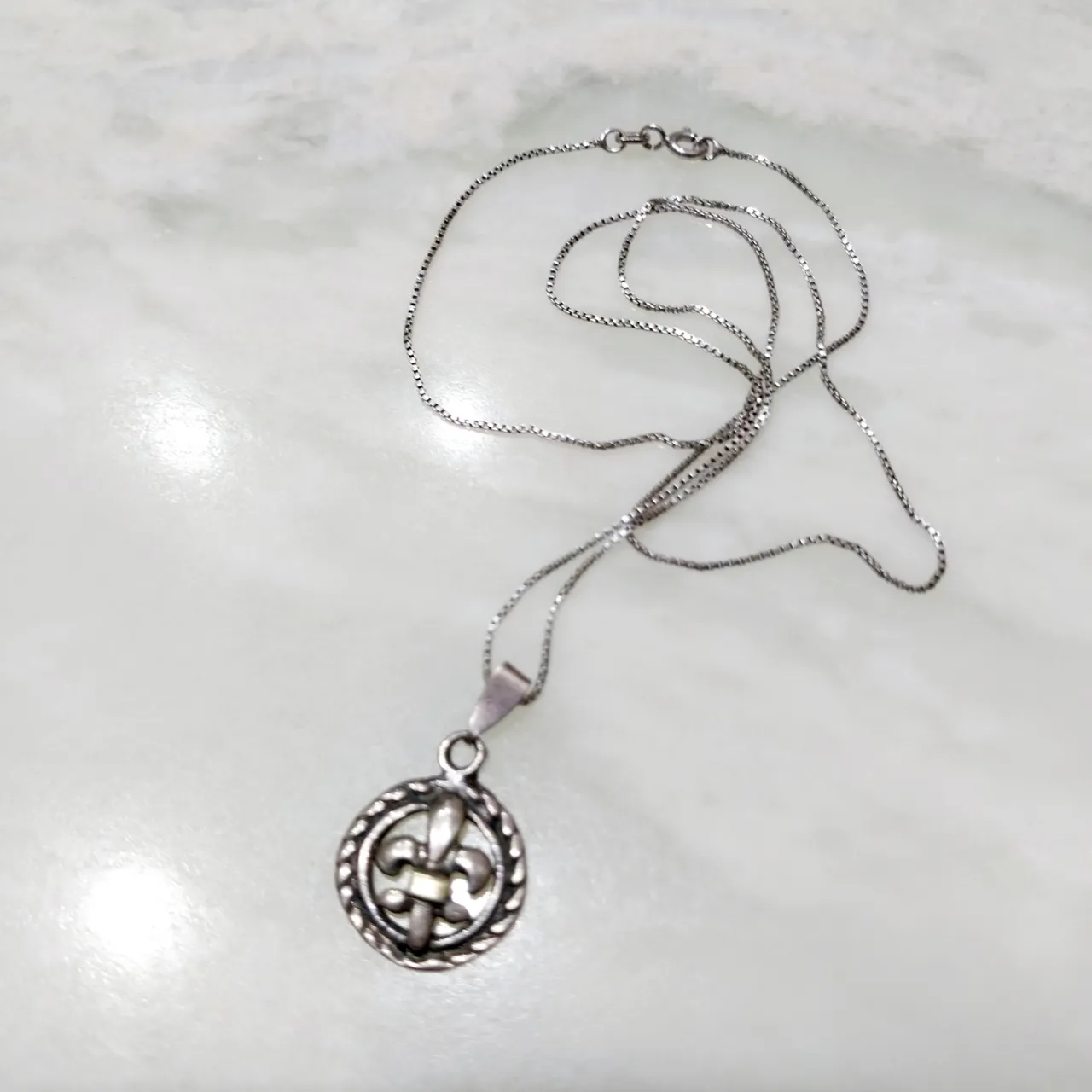 Cadena fina de plata 925 con un dije circular que tiene un diseño de flor de lis en el centro, rodeado por un borde trenzado. Se cierra con un broche tipo mosquetón.