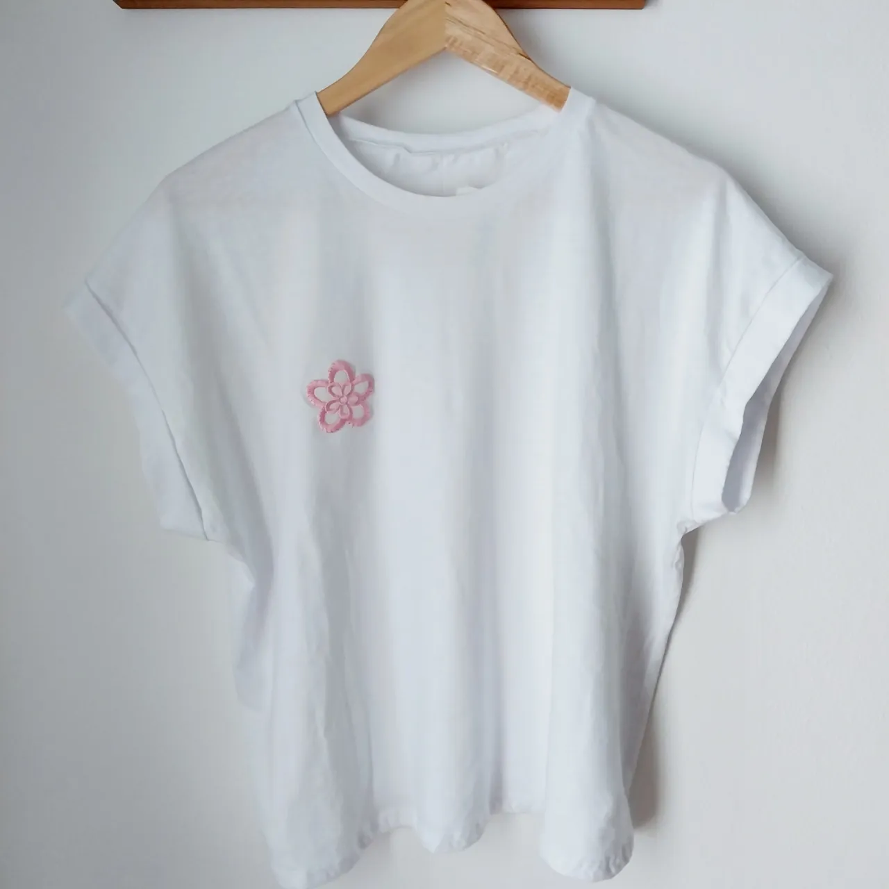 Remera blanca de algodón con un delicado bordado de florcita en color rosa pálido en el frente. Diseño minimalista y fresco, ideal para el día a día. Las mangas son cortas y tienen un dobladillo.