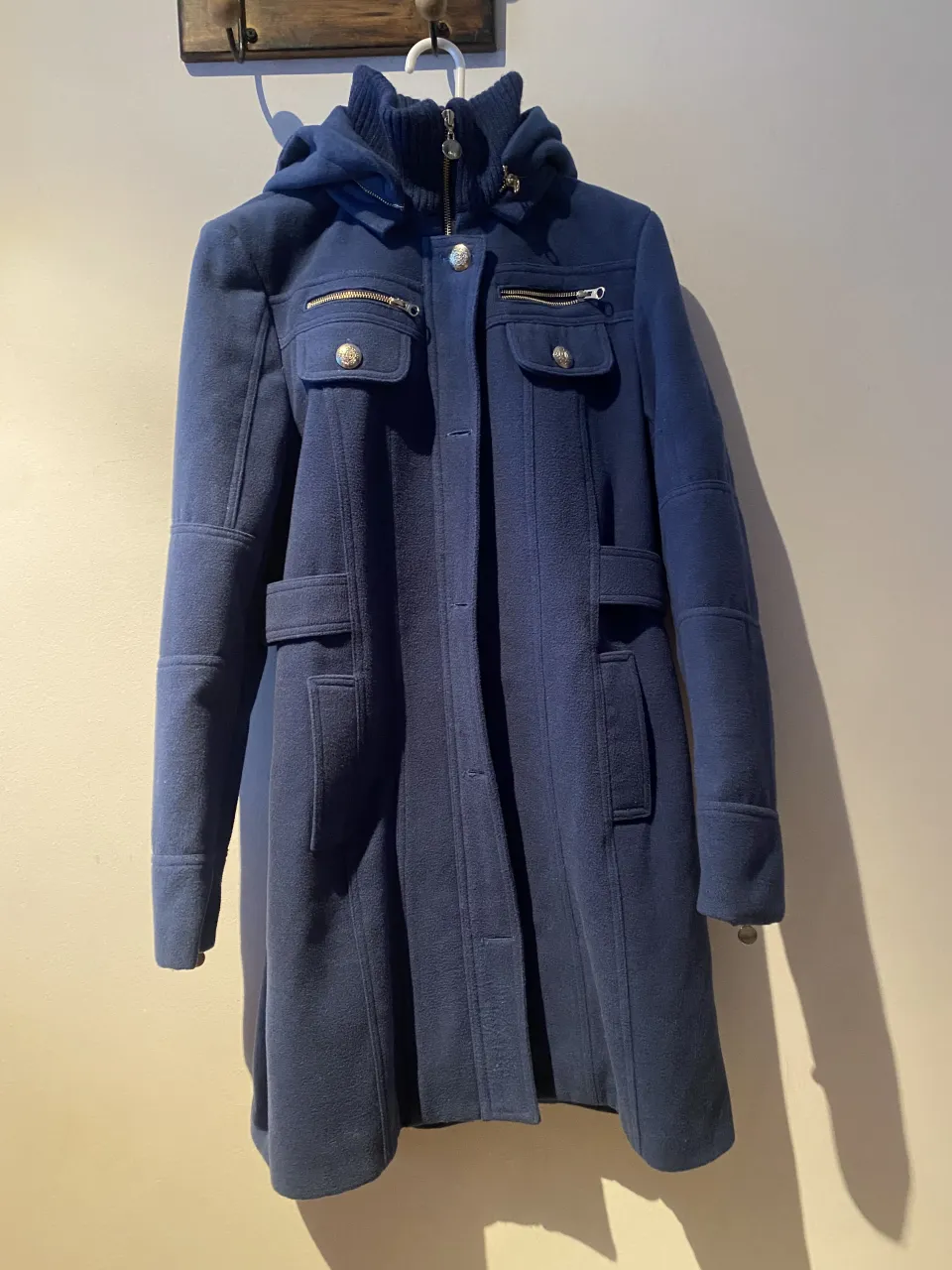 Abrigo azul marino con capucha, cierre y botones. Posee bolsillos con solapa y cierre metálico en el frente. Detalle de cinturón en la espalda con botones.