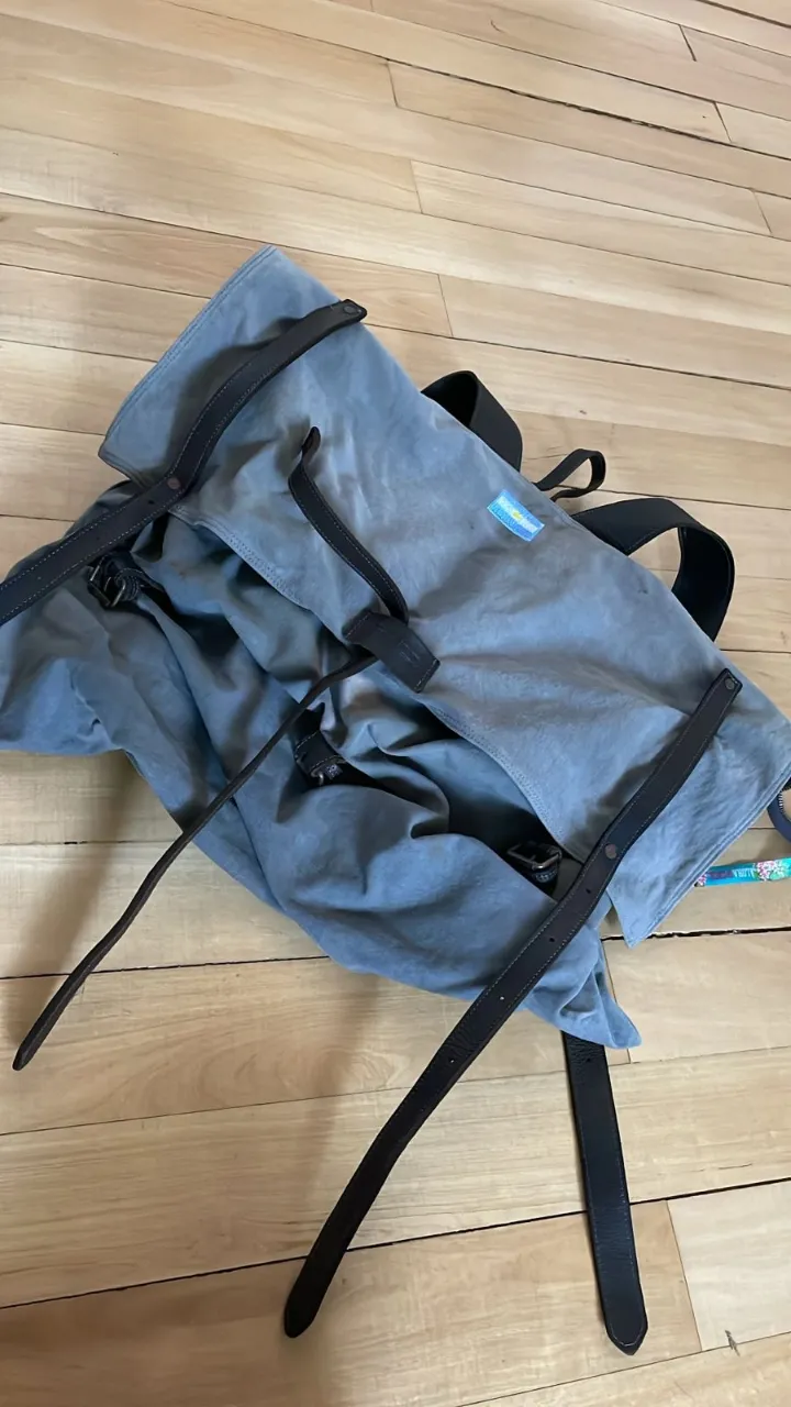 Mochila de lona gris con detalles en cuero negro. Cierre de hebillas y correas ajustables.