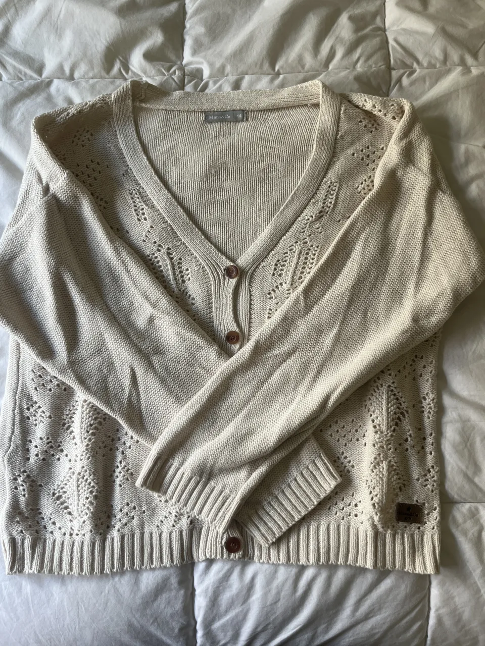 Cardigan de tejido de punto color crudo con botones al frente y detalles de ochos calados en delantero y mangas. Puños y cintura con tejido de canalé.