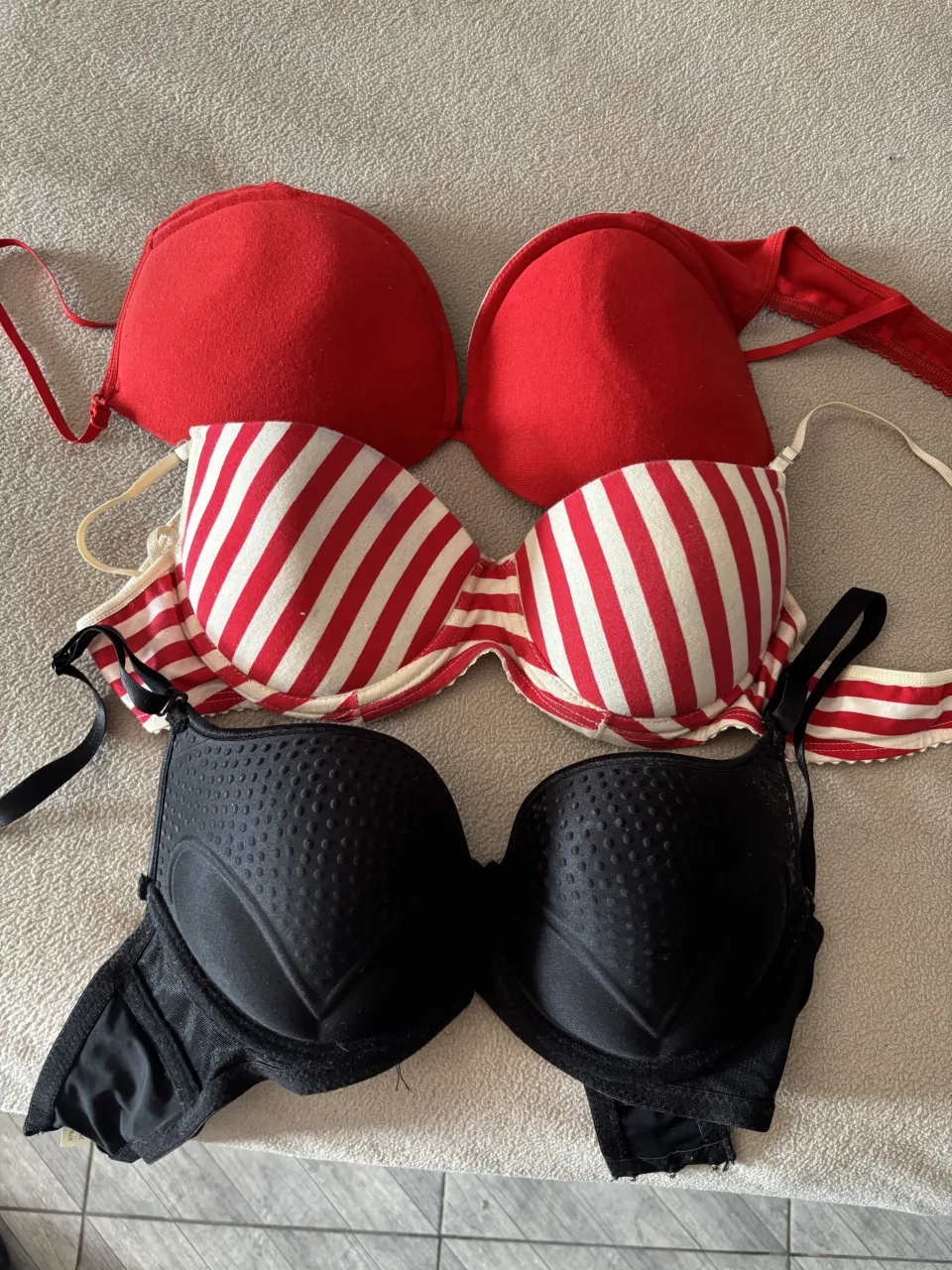 Conjunto de tres corpiños, dos rojos lisos y uno a rayas rojas y blancas. El corpiño negro tiene un detalle de textura con pequeños puntos y un diseño en forma de corazón en las copas. Ideal para el uso diario o para complementar outfits.