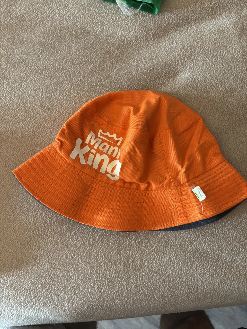 Sombrero tipo "bucket hat" de color naranja vibrante, con un estampado blanco que dice "Man King" y un detalle de corona. Ideal para protegerse del sol con estilo.