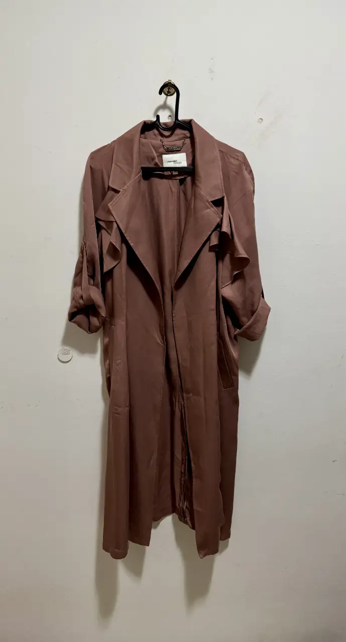 Trench coat largo de color marrón, ideal para un look elegante y sofisticado. Confeccionado en un tejido suave y fluido, este trench presenta un diseño clásico con solapas amplias, mangas largas y un cinturón para ajustar la cintura. Perfecto para usar en cualquier ocasión, desde una salida casual hasta un evento formal.