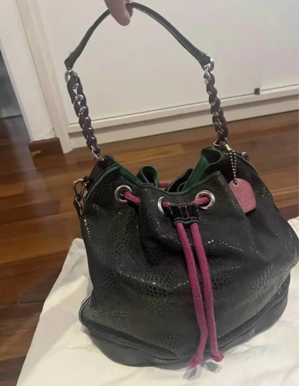Cartera Jackie Smith negra con detalles en rosa y verde. Diseño tipo balde con manija de cadena y correa larga desmontable. Incluye un dije de corazón y correa para llaves. 