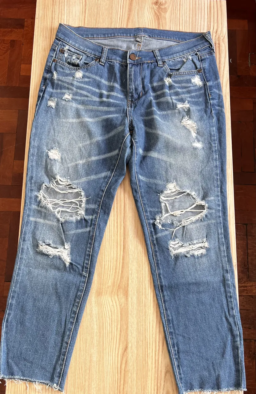 Jeans de denim azul con roturas y desgastes en la parte delantera. Los bordes inferiores de los pantalones están desflecados.