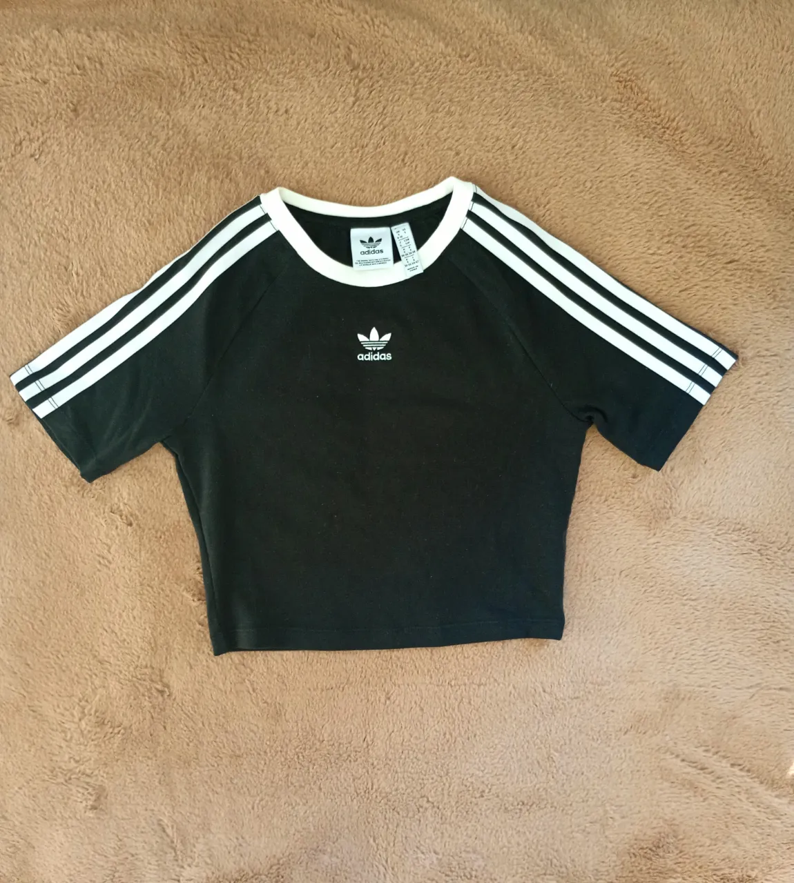 Remera negra corta con cuello redondo y mangas cortas. Posee tres tiras blancas en cada manga y el logo de Adidas en blanco en el frente.