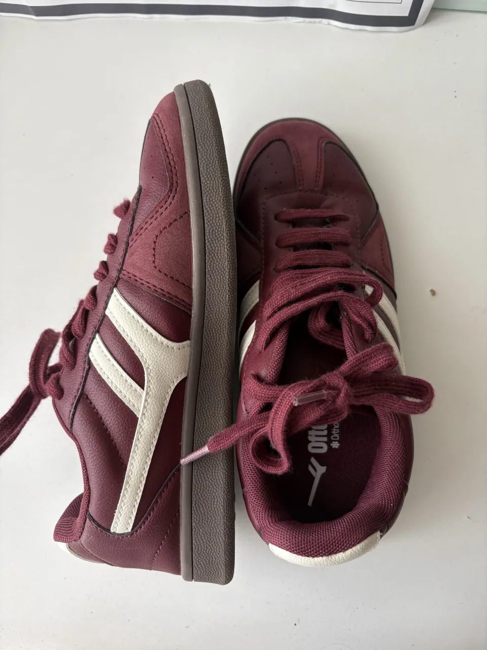 Zapatillas deportivas color bordó con detalles blancos en los laterales y cordones bordó. La suela es de goma color marrón.