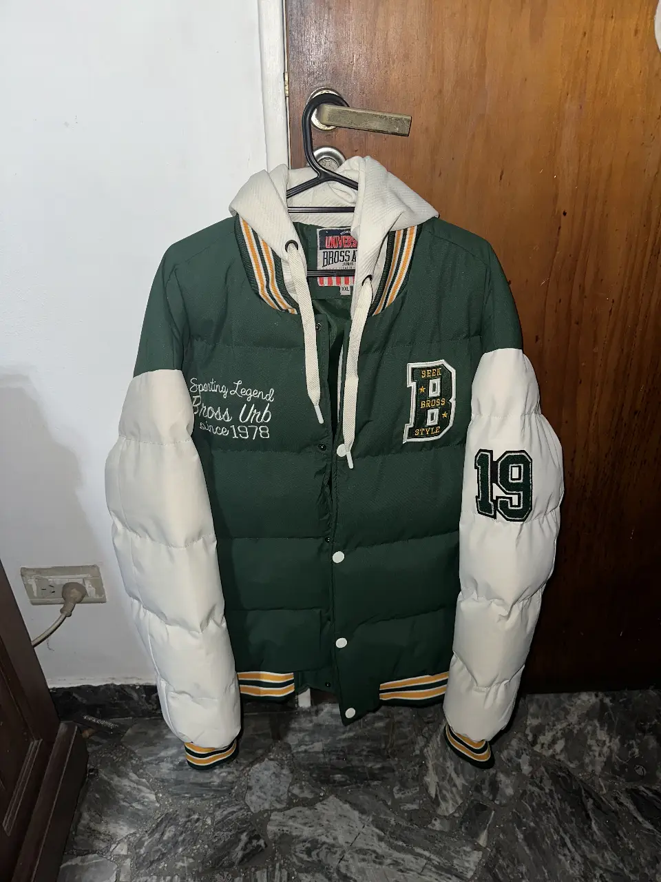 Campera bomber verde con mangas blancas acolchadas, capucha y detalles en amarillo y negro. Estilo colegial con parches y número 19 en la manga. Ideal para un look casual y abrigado.