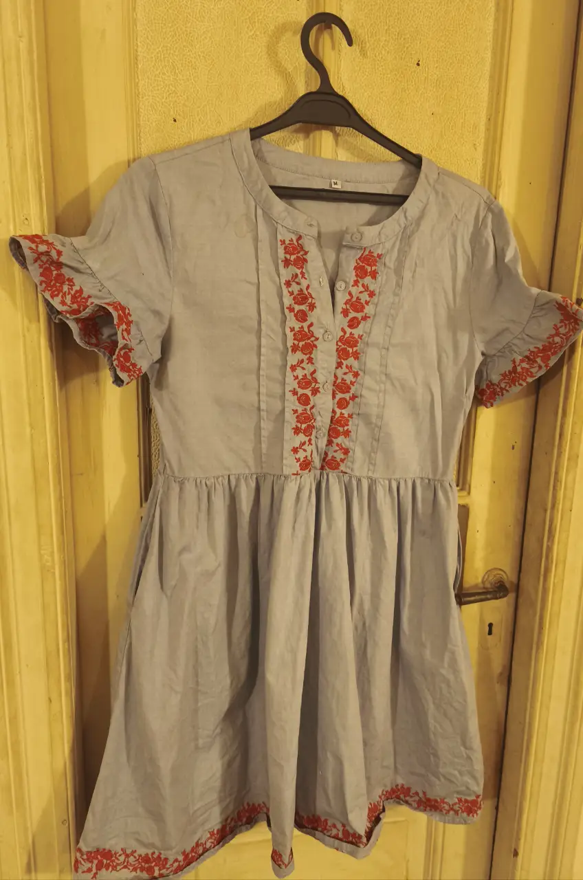 Vestido mini gris con bordado floral rojo en el cuello y mangas. Ideal para un look fresco y casual, con bolsillos.