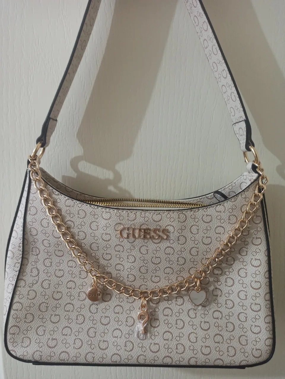 Bolso Guess súper canchero, ideal para el día a día o para darle un toque chic a tu outfit. Tiene un diseño estampado con el logo de la marca y detalles de cadena dorada con charms que le dan un toque único. Es espacioso por dentro y súper práctico.