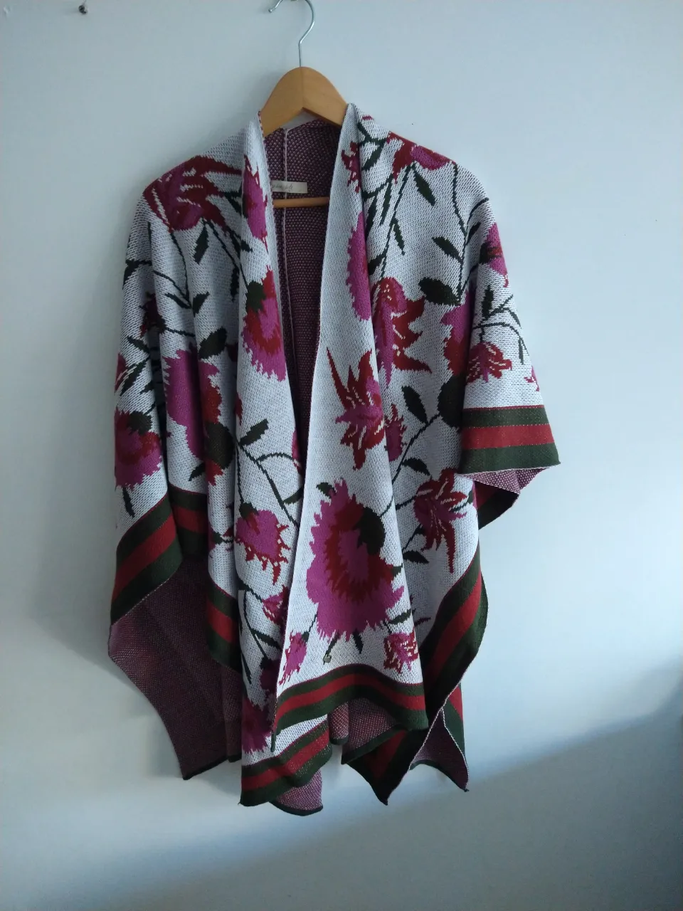 Poncho tejido con estampa floral en tonos fucsia y verde sobre fondo blanco. Posee bordes con franjas horizontales en verde y rojo.