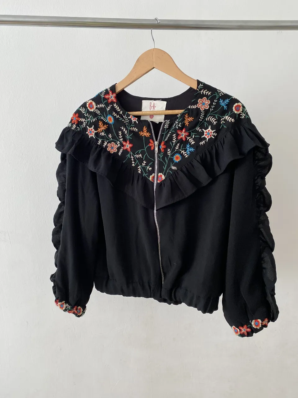Blusa negra con bordado floral multicolor en el cuello y puños. Detalles de volantes y cierre frontal. Ideal para un look bohemio y elegante.