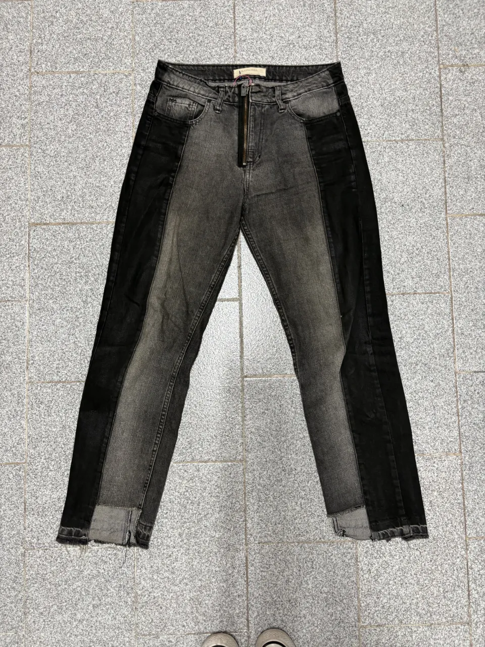 Jeans de corte recto con un diseño moderno y audaz, combinando paneles de denim en diferentes tonos de gris y negro. Poseen cierre metálico al frente y un acabado desflecado en los bajos, dándole un toque canchero y actual. Ideales para un look urbano y descontracturado.