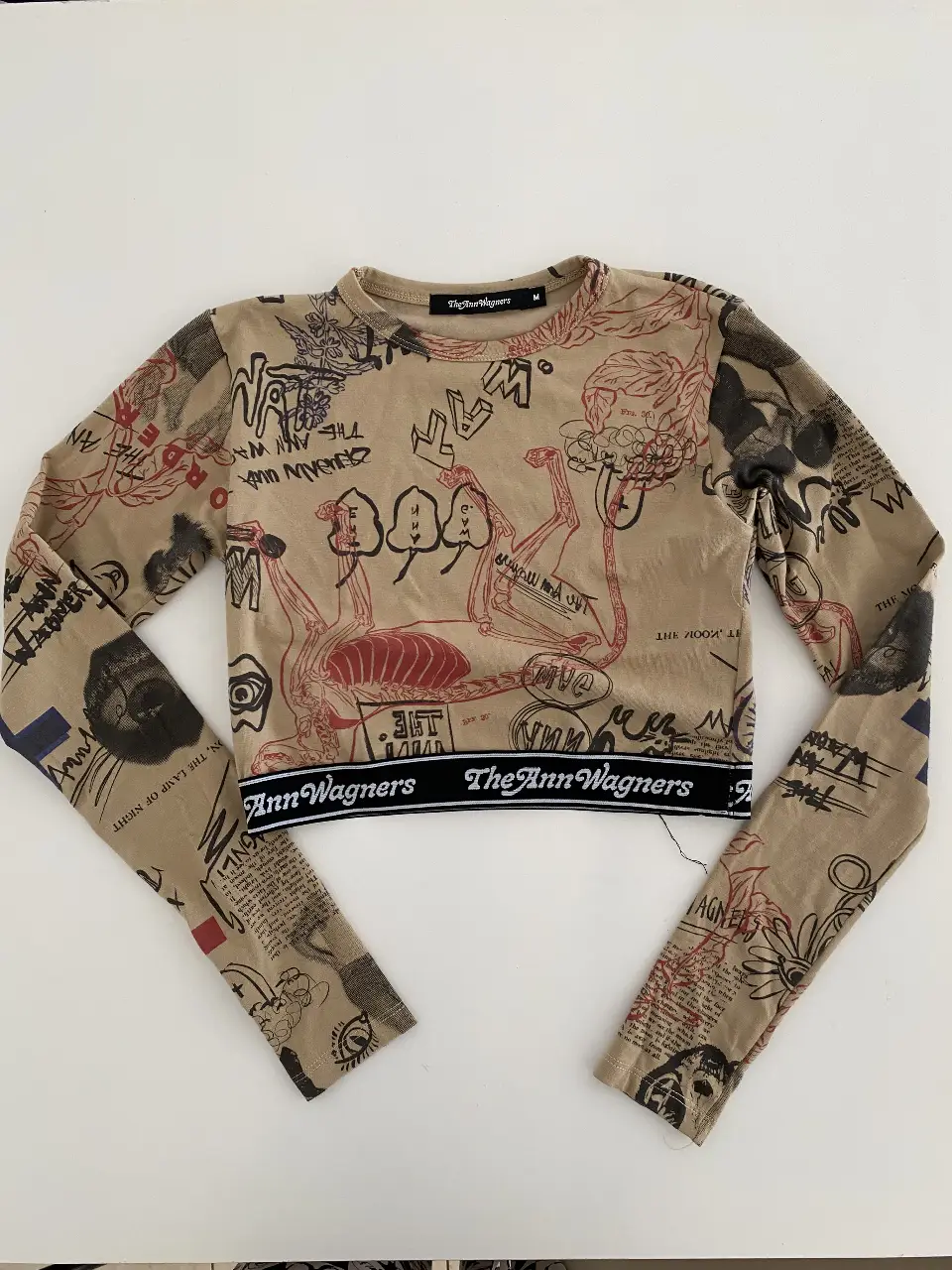 Top manga larga crop con estampado artístico. Diseño con detalles de figuras y texto en negro y rojo sobre fondo crudo. Cintura con el logo de la marca en negro sobre blanco.
