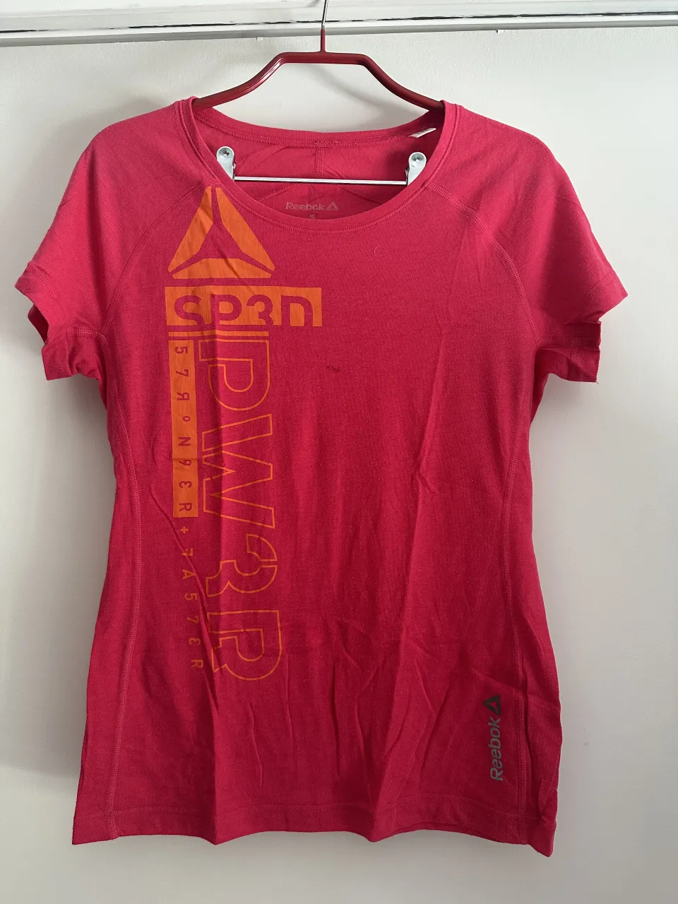 Remera deportiva Reebok talle S, color rosa vibrante con estampa "SP3D" en naranja y gris. Ideal para entrenamientos o uso casual.