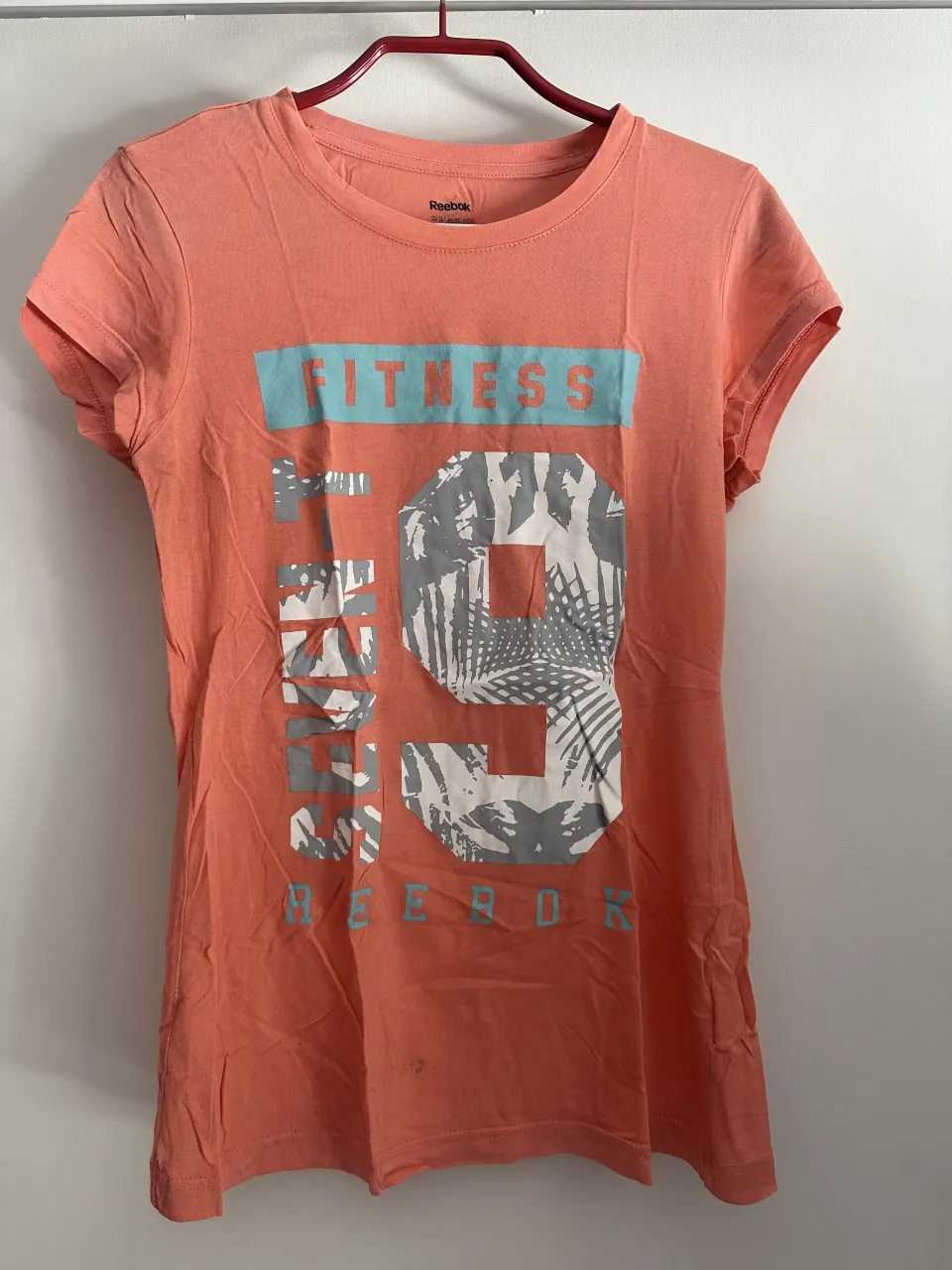 Remera deportiva color salmón de Reebok, con estampa "FITNESS 19" en el frente. Ideal para entrenar o para un look casual y cómodo.