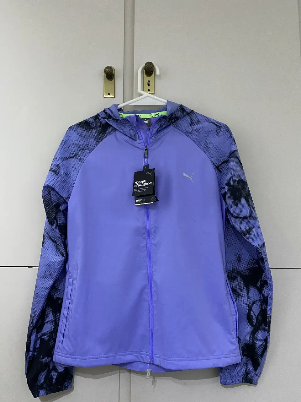 Campera deportiva Puma para mujer, con cierre y capucha. Diseño en color lila con mangas en estampado negro y lila. Ideal para running y actividades al aire libre.
