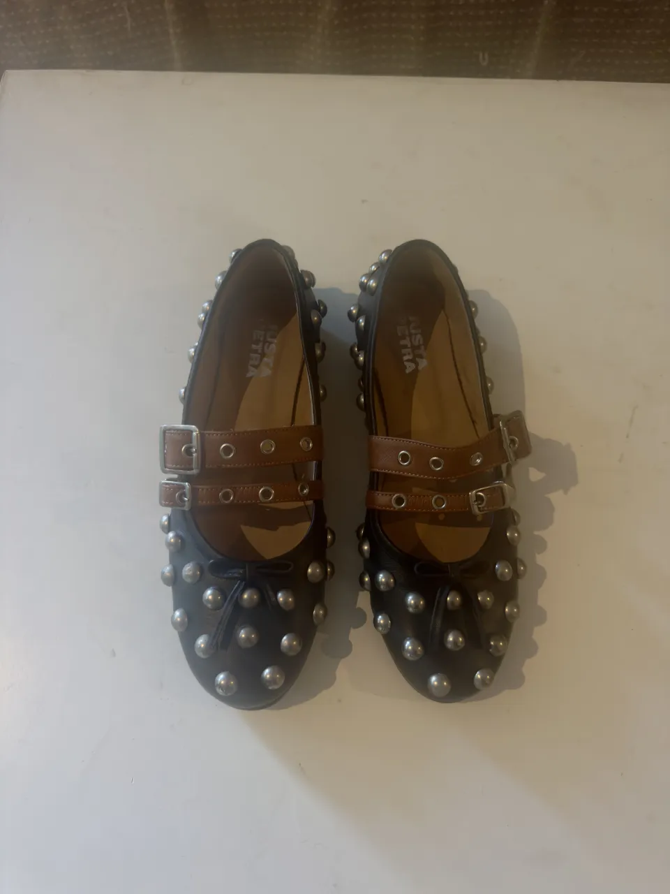 Zapatos planos negros con tachas plateadas y tiras de cuero marrón con hebillas. ¡Un toque rockero y canchero para tu look!