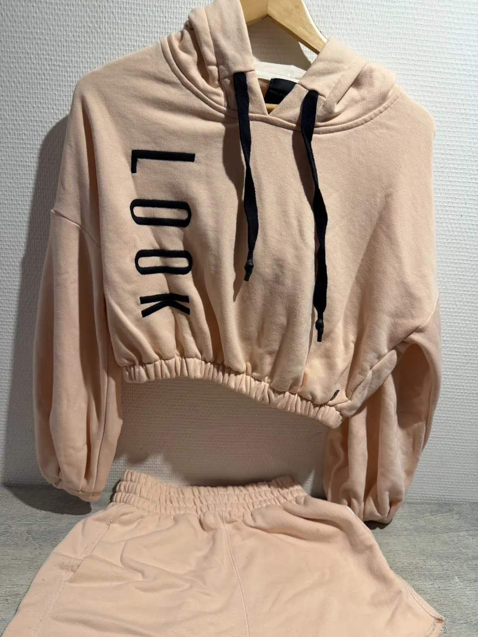 Conjunto de short y buzo crop top color salmón. El buzo es de manga larga, con capucha y cordones negros. Tiene la palabra "LOOK" estampada en negro en el frente. El short es tiro alto con cintura elastizada y bolsillos laterales. El conjunto tiene un estilo casual y moderno, ideal para el día a día o para un look relajado.