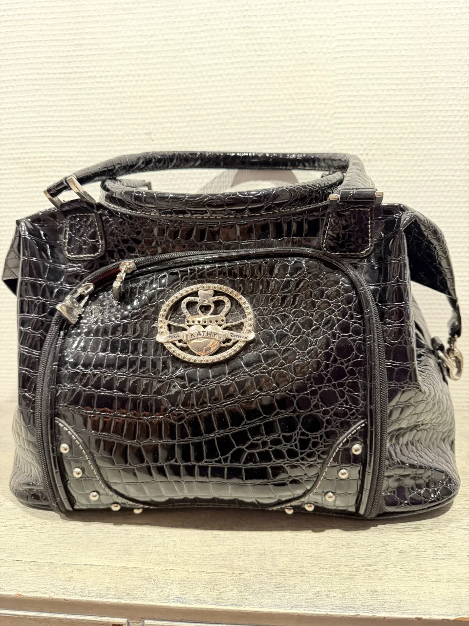 Bolso negro de cuero sintético con textura de cocodrilo, súper brillante y llamativo. Tiene un detalle de aplique metálico con un corazón y coronas en la parte frontal, y tachas plateadas en la base. El interior es de color rosa vibrante y cuenta con un bolsillo interno y otros para collates. Ideal para darle un toque glamoroso a cualquier outfit. Nuevo