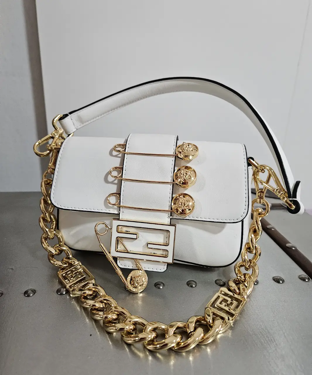 Cartera blanca con detalles dorados y cadena gruesa. Diseño con apliques de broches y cierre con logo.