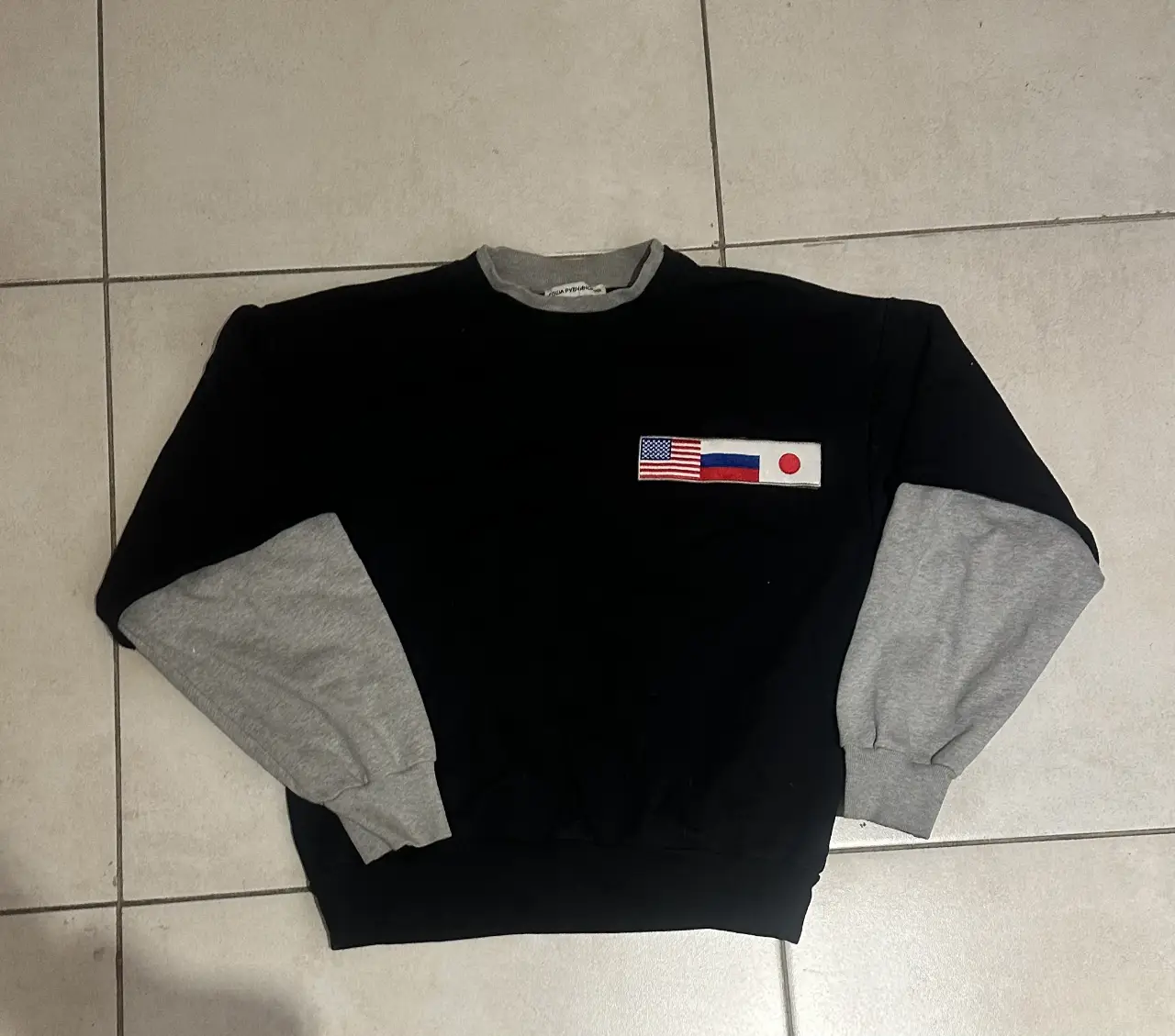 Buzo original de la marca del diseñador ruso Gosha Rubchinsky de cuello redondo negro con mangas grises. Detalles de parches con banderas de Estados Unidos, Rusia y Japón. Cuello y puños en gris. Estilo vintage.