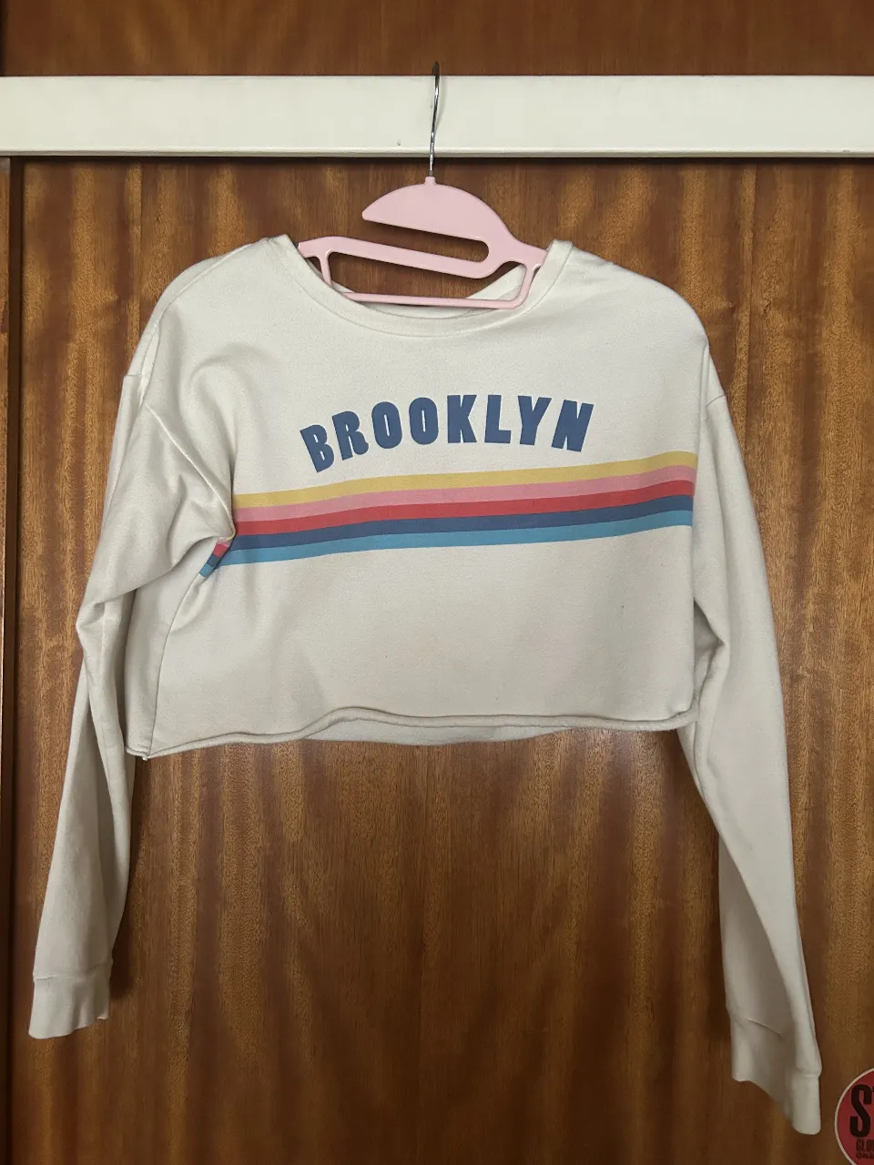 Buzo crop top blanca con estampa de "BROOKLYN" en letras azules y una franja de colores arcoíris debajo. Manga larga y corte en la parte inferior. Ideal para un look casual y moderno.
