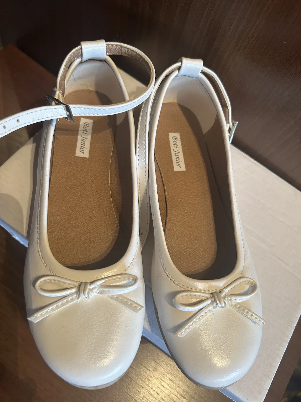 Zapatitos blancos para nena, estilo bailarina, con moño y tira para abrochar en el tobillo. Perfectos para cualquier ocasión especial.