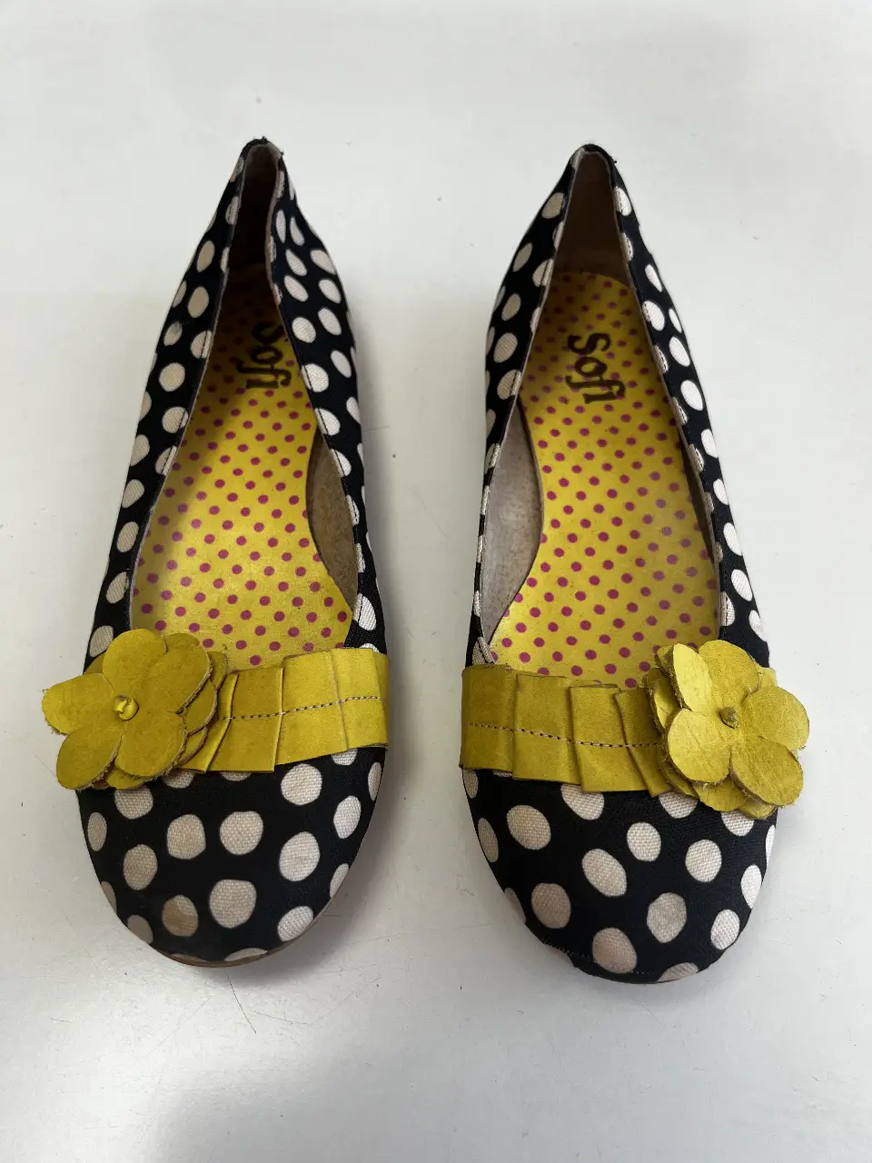 Ballerinas de mujer con estampado de lunares blancos sobre fondo negro. Detalles en amarillo, incluyendo una cinta y una flor decorativa. Interior amarillo con lunares rosas.