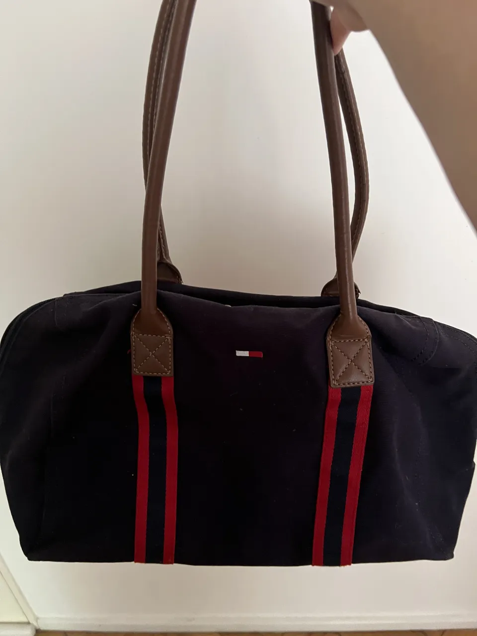 Bolso de tela azul marino con dos tiras de cinta roja y azul a los costados. Tiene dos asas de cuero marrón y un logo de Tommy Hilfiger en la parte frontal. El interior es de tela negra con un bolsillo.