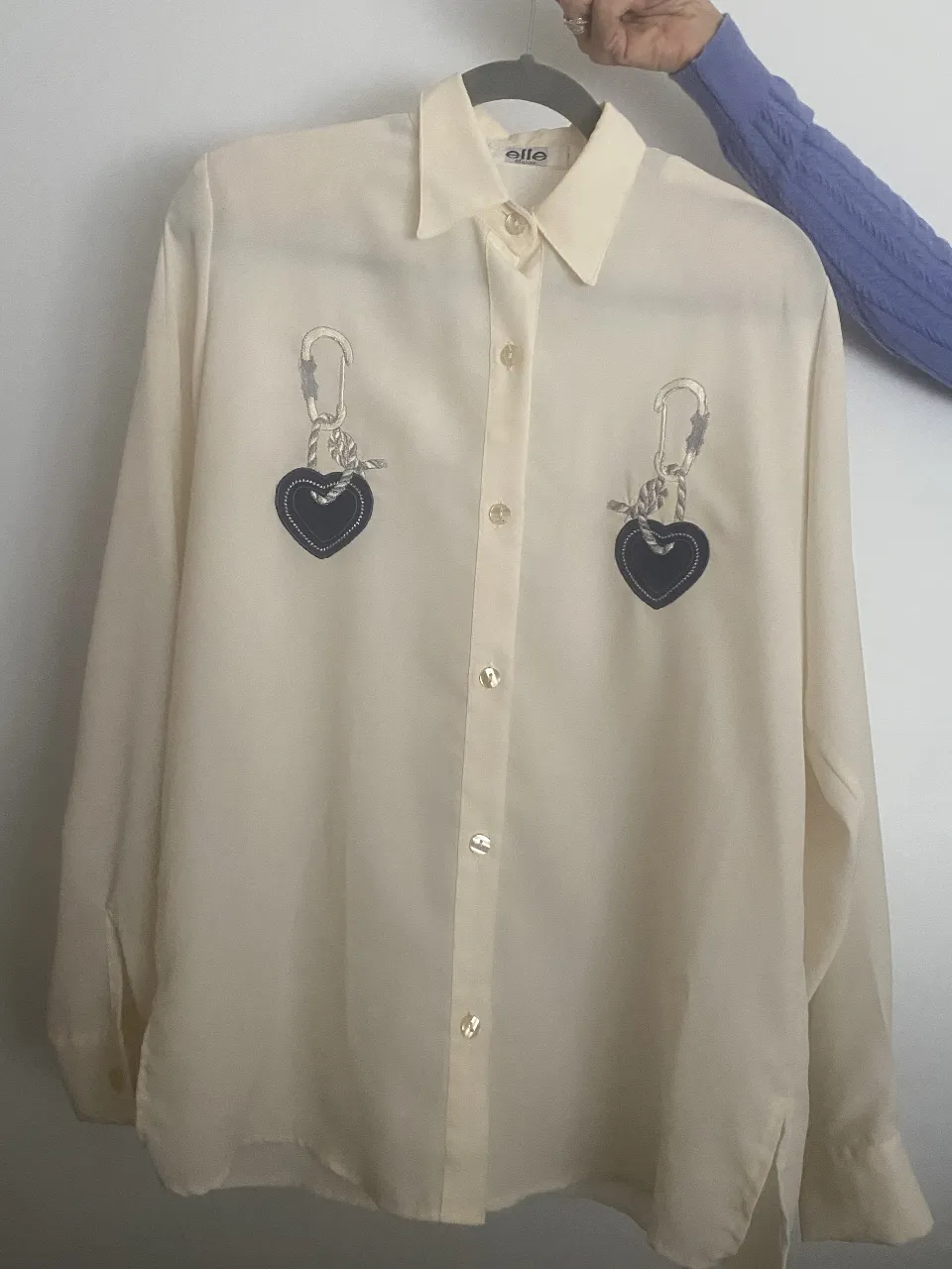 Camisa de seda color crema con bordados de corazones y mosquetones en el pecho. Un diseño único y original para destacar tu estilo.