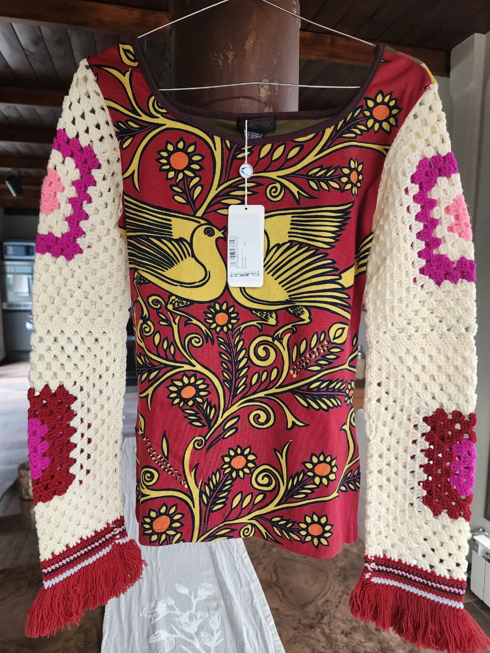 Remera manga larga con estampa de pájaros y flores en el frente. Las mangas son de crochet color crudo con detalles de cuadrados en fucsia y rosa. Posee puños de tejido con flecos en color rojo.