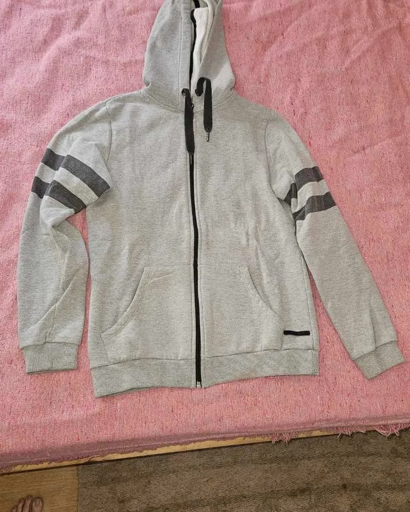 Buzo gris con capucha, cierre completo y detalles de franjas en las mangas. Ideal para un look casual y cómodo.