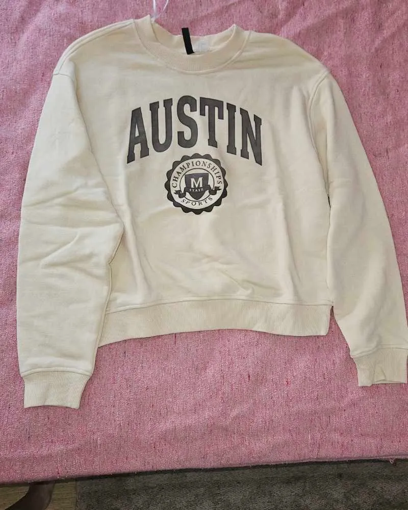 Buzo de algodón color crema con estampa "Austin Championships Sports" en gris oscuro. Diseño oversize y corte cropped, ideal para un look casual y moderno.