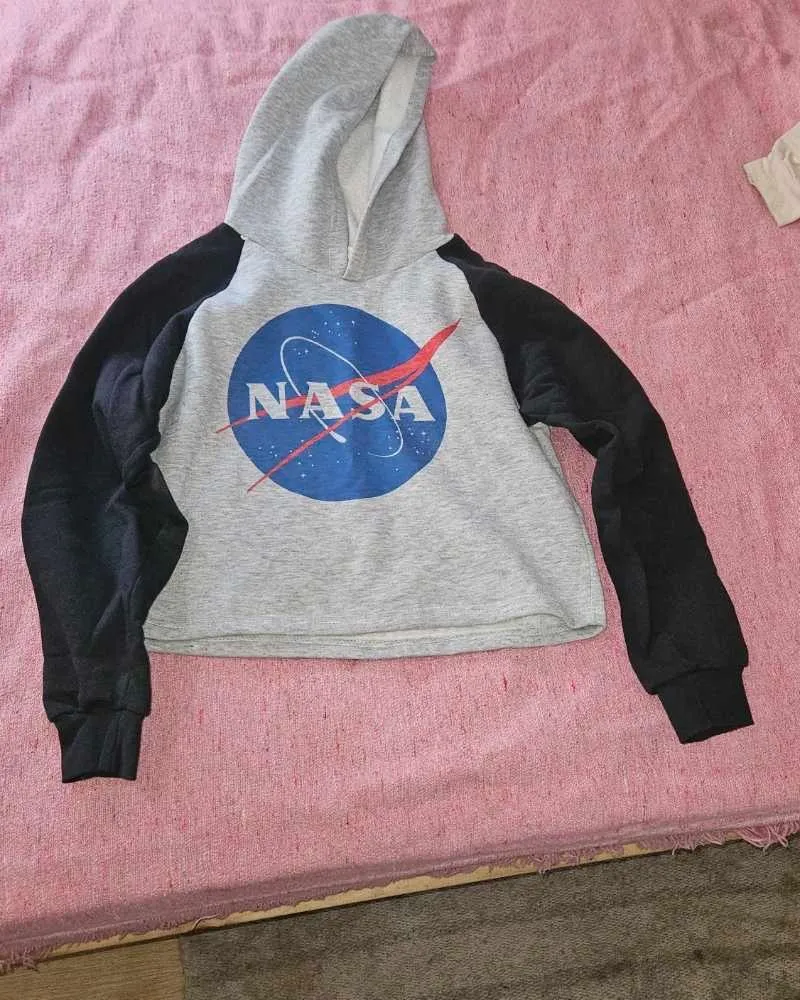Buzo crop top con capucha y mangas largas, gris con mangas negras. Estampado del logo de la NASA en el frente. Ideal para un look casual y moderno.