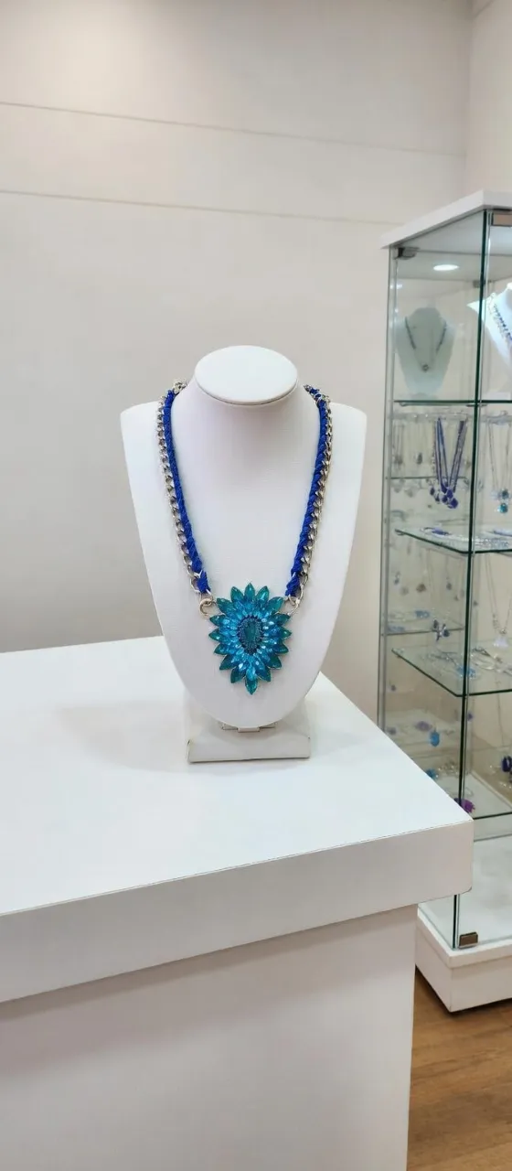 Collar con cadena doble, una de eslabones plateados y otra de eslabones plateados entrelazados con hilo azul. Posee un dije central en forma de flor con pétalos celestes y un centro con piedras turquesas.